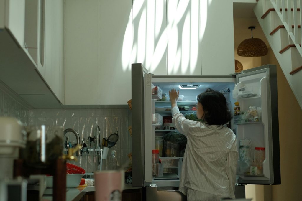 A woman in pajamas checks the refrigerator in a warmly lit cozy kitchen.